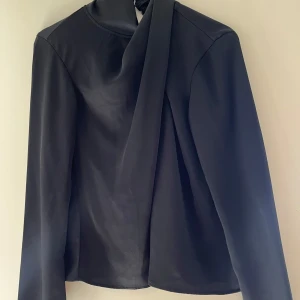 Svart blus med omlotthals  från H&M - Svart långärmad blus från H&M med elegant omlott vid halsen. Toppens material är slätt och har en stilren, rak passform med vida armar med slits. Använd endast en gång💞