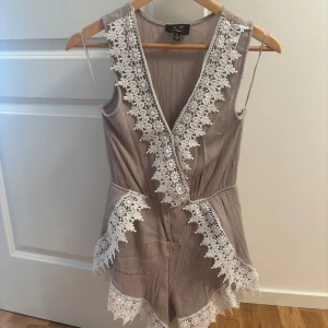 Beige jumpsuit med spets från ICE - Supersnygg beige jumpsuit med omlottkänsla från ICE Fashion. De har vita spetsdetaljer längs kanterna och v-ringad front. Materialet är lätt och luftigt, perfekt för varma dagar. Sitter skönt i midjan och har en feminin vibe med volangliknande spets.