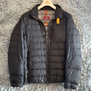 Svart lättviktsdunjacka Parajumpers M - Svart quiltad lättviktsdunjacka från Parajumpers i storlek M. Jackan har dragkedja framtill, ståkrage och diskret gul detalj vid halsen. Tillverkad i syntetmaterial och dun, perfekt för kyliga dagar. Klassisk och stilren design med två sidofickor.