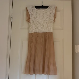 Vit och beige spetsklänning - Supersöt kortklänning med vit blommig spets upptill och plisserad beige kjol. Ärmlös modell med volangdetaljer vid axlarna. Perfekt för sommarens alla tillfällen och ger en romantisk vibe.