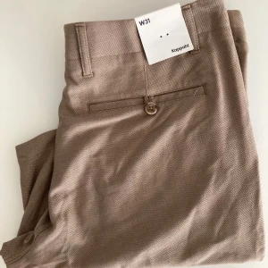Beige kostymbyxor - Snygga beige kostymbyxor från Kappahl i klassisk modell med bälteshällor, knappstängning och diskret struktur i tyget. Byxorna har fickor fram och bak samt stilren passform, perfekta till skjorta eller t-shirt.
