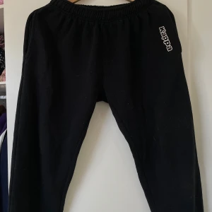 Svarta mjukisbyxor från Kappa - Svarta mjukisbyxor från Kappa med resår i midjan och logga på vänster höft. Byxorna har en avslappnad passform och är perfekta för chill eller träning. Tillverkade i mjukt material för extra komfort. Står storlek S men passar bra även på M. Stretchiga byxor