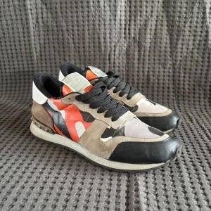 Valentino Rockrunner sneakers i kamouflage - Snygga Valentino Rockrunner sneakers med ett unikt kamouflagemönster i orange, svart, grått och vitt. Skorna har en klassisk snörning och är tillverkade i en kombination av skinn och syntetmaterial. Perfekta för en trendig och avslappnad stil. De är slitna det är anledningen till varför det är så bra pris!