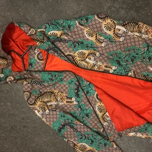 Gucci vindjacka med tigerprint - Fet vindjacka från Gucci med ikoniskt tigerprint, gröna palmblad och GG-mönster på brun bas. Jackan har huva, dragkedja och insida i knalligt orange. Perfekt statement-plagg för dig som vill sticka ut. ( sällsynt )