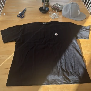 Svart Moncler t-shirt med logga - Svart t-shirt från Moncler med klassisk rund hals och korta ärmar. På bröstet sitter Moncler märket. Säljer pga för stor och den är i helt nytt skick - 10/10.