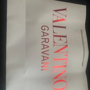 Vit shoppingväska från Valentino Garavani - Säljer en stor och stilren shoppingväska från Valentino Garavani i vit färg med glansigt röd logga framtill. Väskan har rejäla handtag i vitt snöre och är perfekt för dig som vill bära med dig mycket och samtidigt se lyxig ut.