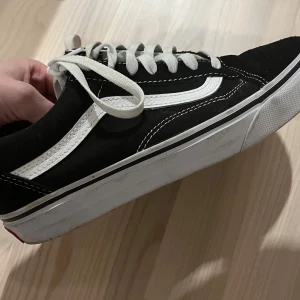 Svarta Vans Old Skool sneakers - Klassiska Vans Old Skool sneakers i svart med vit sidorand och vita snören. Skorna har en låg profil, platt vit sula och tydliga kontrastsömmar. Ovandelen är i mocka och canvas, vilket ger en snygg och tidlös look. Perfekta för dig som gillar streetstyle.