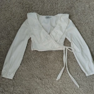 Vit omlottopp med volang från NA-KD - Säljer en vit omlottopp från NA-KD med långa ärmar och volangkant längs v-ringningen. Toppen är croppad och knyts i sidan för en snygg passform. Tillverkad i bomull och har knappar vid ärmsluten. Storlek 34!