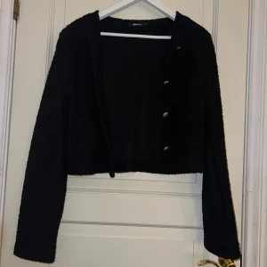 Svart kofta från Gina Tricot - Säljer en svart, croppad kofta från Gina Tricot med långa ärmar och snygga knappar framtill. Koftan har en mjuk, stickad känsla och passar perfekt till lager-på-lager-stil. Enkel att matcha med både jeans och kjol.