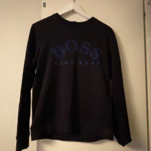 Svart tröja från Hugo Boss - Svart långärmad tröja från Hugo Boss med blå logga tryckt framtill. Klassisk rund halsringning och mjukt material som känns skönt mot huden. Perfekt för dig som gillar stilrena och snygga plagg med tydlig branding.