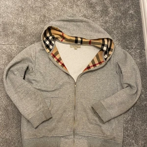 Burberry Hoodie  - Säljer en grå hoodie från Burberry i bomull med klassiskt rutigt foder i huvan. Tröjan har dragkedja framtill, två fickor och ribbade muddar. Perfekt för dig som vill ha en stilren och lyxig vardagslook. Bra skick endast smutsig(tvättas självklart innan den skickas) priset är absolut inte hugget i sten 
