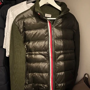 Olivgrön dunväst från Moncler - Snygg olivgrön dunväst från Moncler med quiltad front och stickade ärmar. Västen har dragkedja med röd och vit detalj längs kanten och hög krage. Perfekt för lager-på-lager och ger en sportig vibe.