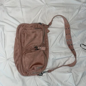 Rosa axelväska från Gina Tricot - Cool rosa axelväska från Gina Tricot i mockaimitation med bred axelrem dekorerad med silvriga nitar. Väskan har flera fack med dragkedja framtill och är perfekt för att få plats med allt du behöver. Snygg och praktisk design som sticker ut.