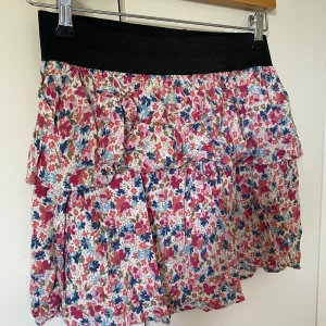 Blommig volangkjol från Gina Tricot - Supersöt kort kjol från Gina Tricot med blommigt mönster i rosa, blått och beige. Kjolen har bred svart resår i midjan och flera volanger som ger en luftig och lekfull känsla. Perfekt för dig som gillar färg och vill sticka ut lite extra.