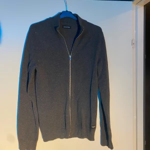 Mörkgrå kofta från Jack & Jones - Mörkgrå kofta från Jack & Jones med hel dragkedja framtill och ribbad krage. Tröjan är långärmad och har en stilren, enkel design som passar perfekt till lager-på-lager. Materialet är mjukt och stickat för extra komfort.