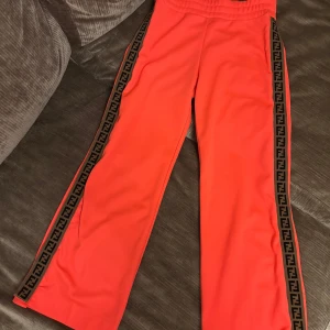 Fendi strl XS - Snygga korallfärgade byxor från Fendi med svarta/bruna logoband längs sidorna. Byxorna har elastisk midja, raka ben och tryckknappar längs hela sidorna. Tillverkade i mjuk polyester för en sportig vibe. Det är storlek 12A i barn/ungdom som passar mig som är XS och 1,56 m lång. Hittar inget att anmärka på!