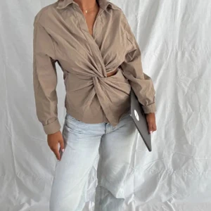Beige skjorta med knyt framtill - Snygg beige skjorta i bomull med lång ärm och krage. Skjortan har en unik knyt-detalj framtill som ger en cool twist och lite croppad känsla. Perfekt att styla med jeans för en trendig look.