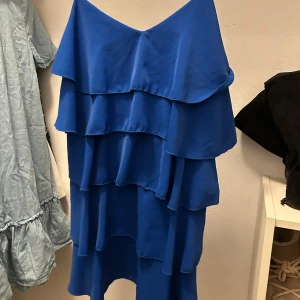 Blå volangklänning Zebra a Pua - Supersnygg blå klänning från Zebra a Pua med flera lager volanger och v-ringad hals. Klänningen har en luftig passform och är ärmlös, perfekt för varma dagar. Tillverkad i ett lätt och mjukt material som ger ett härligt fall.