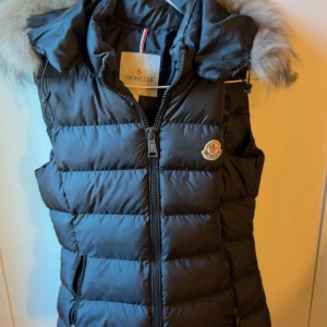 Svart dunväst med päls - Svart dunväst från Moncler med fluffig päls runt huvan och quiltad design. Västen har dragkedja framtill, två fickor och klassisk Moncler-logga på bröstet. Perfekt för kyliga dagar när du vill ha både stil och värme.( fick som present! )