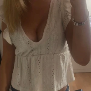 Vit blus med volang  - Supersöt vit blus från Pull&Bear med broderade detaljer och volang vid axlarna. Djup v-ringning och resår i midjan ger en snygg siluett. Blusen är ärmlös och har ett luftigt, lätt material.