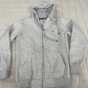 Grå hoodie från Tommy Hilfiger - Snygg ljusgrå hoodie från Tommy Hilfiger med dragkedja, huva med snörning och två stora fickor framtill. Mjuk insida och diskret logga på bröstet. Perfekt för chill dagar eller när du vill ha en clean och enkel stil.