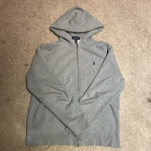 Grå hoodie från Polo Ralph Lauren XL - (Pris kan alltid diskuteras).              Klassisk grå hoodie från Polo Ralph Lauren i storlek XL. Tröjan har dragkedja, huva och två fickor framtill. Broderad marinblå logga på bröstet. Tillverkad i mjuk bomullsmix, perfekt för chill dagar.