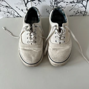 Vita sneakers från Polo Ralph Lauren - Snygga vita sneakers från Polo Ralph Lauren med klassisk design. Skorna har ovandel i bomull, detaljer i brunt skinn vid hälen och rutigt foder i grönt och svart. Broderad logga på sidan och snörning framtill. Perfekta för en clean och stilren look.