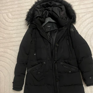 Svart lång dunjacka med fuskpäls - Säljer en svart lång dunjacka från Ragwear med huva och fuskpäls. Jackan har dragkedja, knappar och flera fickor framtill. Perfekt för kalla dagar med sin varma vaddering och snygga detaljer.