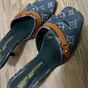 Louis Vuitton denim sandaler med klack - Unika sandaler från Louis Vuitton i blått denim med ikoniskt monogrammönster. Snygga bruna skinn-detaljer och guldfärgade nitar. Klack och öppen tå med fyrkantig form ger en cool vibe. Perfekta för dig som vill sticka ut med designerstil.