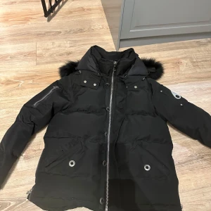 Moose Knuckles dunjacka - Säljer en svart dunjacka från Moose Knuckles med pälskantad huva och coola metalldetaljer på ärmen. Jackan har dragkedja, knappar och flera fickor med snygga detaljer. Perfekt för kalla dagar och riktigt street i stilen. Kom pm för fler bilder