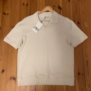 Beige ribbad piké från Zara - Snygg beige piké från Zara med ribbad struktur och klassisk krage, helt ny aldrig använd. Kortärmad modell i mjuk bomull som ger en clean och fräsch look. Perfekt för dig som gillar stilrena plagg med lite extra detaljer.
