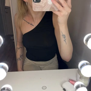 Svart one shoulder topp - Säljer en svart, tight och stilren one shoulder topp med asymmetrisk skärning. Toppen är ärmlös på ena sidan och har en axelrem på andra, vilket ger en snygg och modern look. Perfekt att matcha med både jeans och kjol.