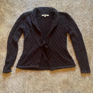 Svart stickad kofta❤️ - Mysig svart stickad kofta från first class med snyggt kabelstickat mönster och knytband i midjan. Koftan har lång ärm och en bred krage som ger en chill och stilren look. Perfekt att slänga på sig när det är lite kyligt ute.