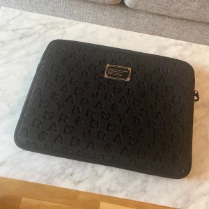 Marc by Marc Jacobs Svart Laptopfodral Datorväska - Snygg svart datorväska från Marc by Marc Jacobs med präglade bokstäver över hela ytan och en metallbricka framtill. Väskan har dragkedja och är tillverkad i mjukt scubamaterial. 