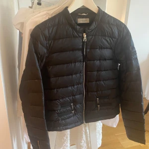 Svart dunjacka från Hunkydory - Svart dunjacka från Hunkydory med stilrena dragkedjor på fickorna och ärmsluten. Jackan har en rund halsringning och är lätt vadderad. Snygg och enkel design som funkar till många olika looks.