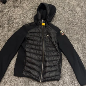 Svart pufferjacka från Parajumpers - Svart pufferjacka från Parajumpers med huva och coolt emblem på ärmen. Jackan har quiltad framsida och rygg, dragkedja framtill och en gul detalj vid halsen. Materialet är syntet och polyester, perfekt för kalla dagar. Snygg och sportig look med långärmade svarta partier på sidor och ärmar.