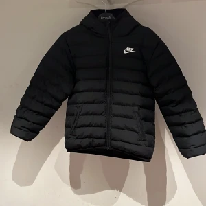 Svart pufferjacka från Nike L - Svart pufferjacka från Nike med huva och vit logga på bröstet. Jackan har dragkedja framtill, två sidofickor och quiltad design. Perfekt för kalla dagar och riktigt snygg streetstil. Priset är kan förhandlas.
