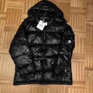 Svart Moncler dunjacka med huva - Svart glansig dunjacka från Moncler med huva och klassisk logga på ärmen. Jackan har dragkedja framtill, elastiska muddar och quiltad design. Perfekt för kalla dagar och riktigt snygg streetstil. Innerfoder med serietidningsdetalj och märkestaggar kvar. Pris kan diskuteras vid snabb affär 