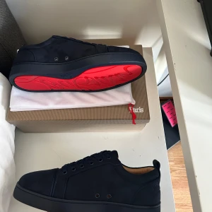 Christian Louboutin mörkblå sneakers - Snygga svarta sneakers från Christian Louboutin med klassisk röd sula. Skorna har ovandel i mocka och skinn, rund tå och svarta snören. Insidan är beige och sulan är platt. Perfekta för dig som vill sticka ut med exklusiv stil.