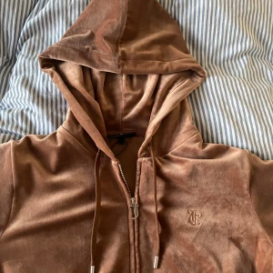 hoodie från Juicy Couture - Säljer en brun hoodie i mjuk velour från Juicy Couture med dragkedja och huva. Tröjan har långa ärmar, fickor framtill och broderad JC-logga på bröstet. Perfekt för dig som gillar en chill och trendig stil. Köpt på NK för 1200kr passar någon som är mellan 160-170cm. Använd någon enstaka gång.