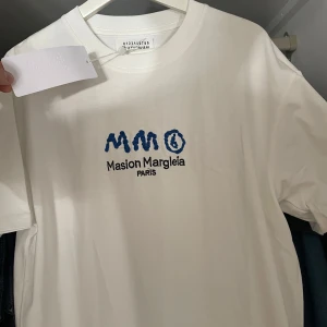 Vit t-shirt Maison Margiela Paris - Vit t-shirt från Maison Margiela Paris med blå broderad logga på bröstet. Klassisk rund halsringning och korta ärmar. Tillverkad i mjuk bomull för en clean och stilren look. Storlek M