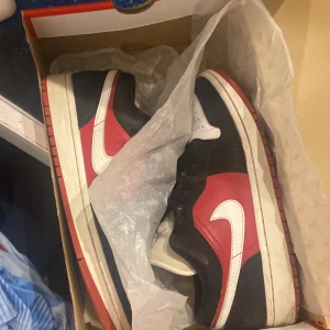 Nike Air Jordan 1 Low Chicago - Säljer ett par Nike Air Jordan 1 Low Chicago med klassisk färgblockning i rött, svart och vitt. Skorna har svart mocka och rött läder på sidorna, vit tåbox och vit swoosh. Svarta skosnören och perforerad tå för extra stil. Perfekta för dig som gillar streetwear och sneakerskultur.