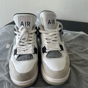 Nike Air Jordan 4 vit/svart/grå - Säljer ett par Nike Air Jordan 4 sneakers i vitt, svart och grått. Skorna har klassisk meshpanel, snörning och detaljer i mocka och skinn. Ikonisk AIR-logga på plösen och hälen. Perfekta för dig som gillar streetwear och vill sticka ut med en tidlös siluett.