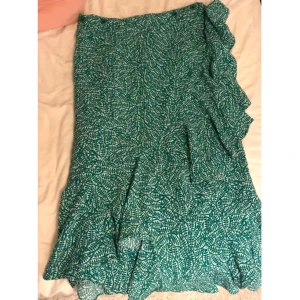 Grön mönstrad kjol - Säljer en kjol från Rodebjer i grönt och vitt med ett snyggt bladmönster. Kjolen har volangdetaljer längs kanten och är i ett lätt, luftigt material som passar perfekt till varmare dagar. Omlottmodell för en extra snygg siluett. (Dyrt märke därav priset)