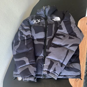 The North Face x KAWS dunjacka camo - Säljer en fet dunjacka från The North Face x KAWS med grått och svart camomönster. Jackan har broderade XX-detaljer, hög krage, dragkedja och två sidofickor. 700-fill dun för max värme och cool streetstyle. Perfekt för kalla dagar.