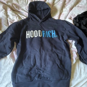 Marinblå HOODRICH hoodie i medium - Säljer en marinblå hoodie från HOODRICH i storlek M.  Tröjan har stor vit och blå logga framtill, huva med snörning och en stor magficka. Det finns även en liten ficka vid armen. 