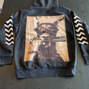 Svart hoodie Bastille Rive Droite - Svart hoodie med stor printad bild av en dobermann på ryggen och texten 'Bastille Rive Droite'. Ärmarna har vita och svarta sicksackmönster. Huva med snörning och stor magficka framtill. Perfekt statement-plagg för dig som gillar streetstyle. Storlek Medium sparsamt använd