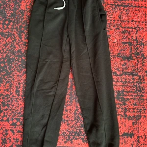 Svarta mjukisbyxor med dragsko - Svarta mjukisbyxor med elastisk midja och vit dragsko. Byxorna har en avslappnad passform och resår vid bensluten. Perfekta för chill eller träning, tillverkade i mjukt material för extra komfort.