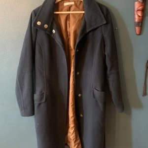 Marinblå lång ullkappa med stora knappar - Stilren svart kappa i ull med klassisk krage och stora dekorativa metallknappar upptill. Fodret är brunt och jackan har två fickor framtill. Perfekt för dig som gillar en tidlös och clean look under kallare månader.