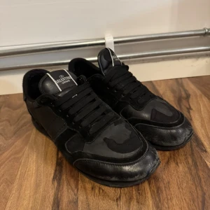 Valentino Garavani svarta sneakers - Svarta sneakers från Valentino Garavani med coola camouflagedetaljer. Skorna har snörning, rund tå och är tillverkade i en mix av skinn och textil. Perfekta för dig som gillar en stilren men edgy look.